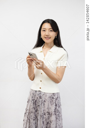 白い壁の前でスマートフォンを持ってカメラ目線で微笑む若い日本人女性 142854607