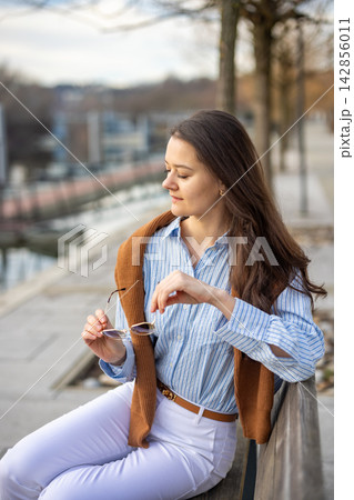 Elegant young woman in old money style sitting on bench near pier in Prague holding sunglasses in early spring. Understated luxury and timeless European femininity concept. Elegant young woman in old money style sitting on bench near pier in Prague holding sunglasses in early spring. Understated luxury and timeless European femininity concept. 142856011