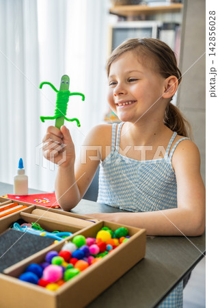 Happy little girl holding handmade craft toy made from wooden stick and pipe cleaners at table. Kids creativity imagination development and learning through play concept. Happy little girl holding handmade craft toy made from wooden stick and pipe cleaners at table. Kids creativity imagination development and learning through play concept. 142856028