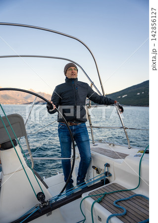 Young man captain standing at the helm and controls a sailboat during a journey by sea in winter time in Montenegro Young man captain standing at the helm and controls a sailboat during a journey by sea in winter time in Montenegro 142856127
