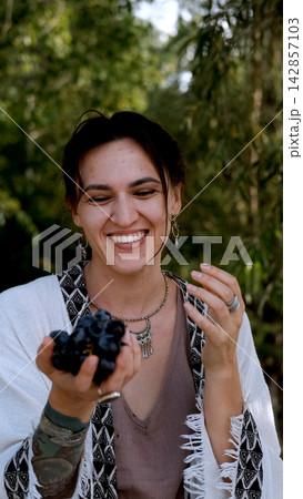 A beautiful brunette in a bohemian style relaxes outdoors in summer against a backdrop of a green forest, holding a large bunch of juicy black grapes A beautiful brunette in a bohemian style relaxes outdoors in summer against a backdrop of a green forest, holding a large bunch of juicy black grapes 142857103