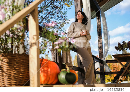 Woman in cozy sweater drinking warm tea on wooden cottage porch with autumn decorations and pumpkins, enjoying sunny morning weather Woman in cozy sweater drinking warm tea on wooden cottage porch with autumn decorations and pumpkins, enjoying sunny morning weather 142858564