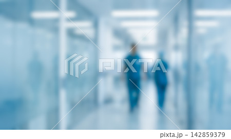 Blurred hallway inside modern hospital featuring male and female medical staff wearing dark scrubs walking past glass walls toward distant bright light 142859379