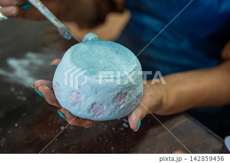 Hands applying glaze to outer surface of handmade ceramic mug with brush. Close up pottery glazing process represents craftsmanship and handmade ceramic production Hands applying glaze to outer surface of handmade ceramic mug with brush. Close up pottery glazing process represents craftsmanship and handmade ceramic production 142859436