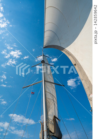 Curved white sail towering above a sailing yacht under a blue sky. Wind powered sailing and freedom on the open sea. Curved white sail towering above a sailing yacht under a blue sky. Wind powered sailing and freedom on the open sea. 142859642