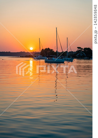 Sailing yachts anchored in a calm bay during a colorful summer sunset over the sea. Peaceful evening, golden hour, coastal travel destination and relaxing sailing lifestyle. 142859848
