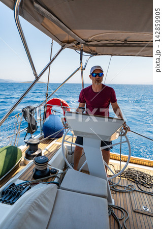 Experienced sailor steering a sailing yacht during an offshore passage at sea. Seamanship, navigation skills, confident boat handling and active cruising lifestyle. Experienced sailor steering a sailing yacht during an offshore passage at sea. Seamanship, navigation skills, confident boat handling and active cruising lifestyle. 142859905