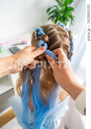 Mother braiding colorful synthetic hair extensions into a young girls hairstyle at home. Creative self expression reflects childhood individuality fashion trends and Generation Alpha style Mother braiding colorful synthetic hair extensions into a young girls hairstyle at home. Creative self expression reflects childhood individuality fashion trends and Generation Alpha style 142860105