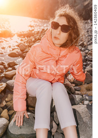 Woman sunset beach sitting in orange puffer jacket on rocks by the sea during golden hour for serene coastal relaxation Woman sunset beach sitting in orange puffer jacket on rocks by the sea during golden hour for serene coastal relaxation 142860409