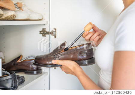 Person removes a wooden shoe tree from a leather shoe near an open wardrobe. Seasonal footwear storage includes maintenance before shoes return to daily wear. Person removes a wooden shoe tree from a leather shoe near an open wardrobe. Seasonal footwear storage includes maintenance before shoes return to daily wear. 142860420