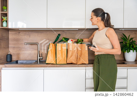 Woman checking grocery delivery with smartphone while unpacking reusable shopping bags in kitchen. Online food ordering concept 142860462