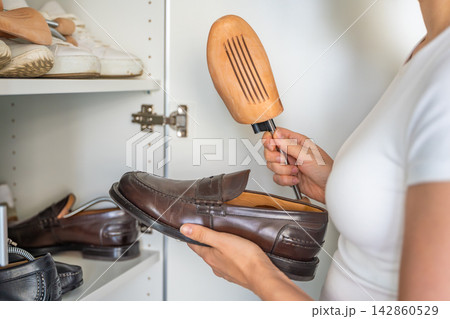 Person holds a leather shoe and a wooden shoe tree near an open wardrobe. Regular shoe care supports seasonal storage and helps retain footwear form. 142860529
