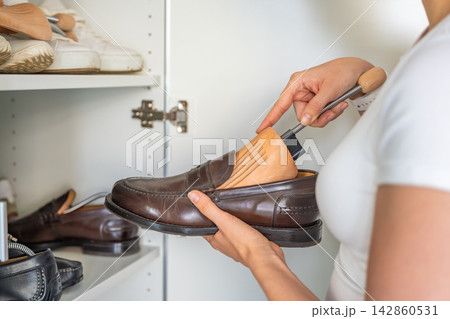 Person inserts a wooden shoe tree into a leather loafer beside a wardrobe. Crease prevention and shape retention improve long term footwear storage. Person inserts a wooden shoe tree into a leather loafer beside a wardrobe. Crease prevention and shape retention improve long term footwear storage. 142860531