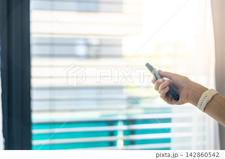 Woman controlling exterior window blinds with a remote control inside a modern apartment. Smart home automation and indoor climate control concept. Woman controlling exterior window blinds with a remote control inside a modern apartment. Smart home automation and indoor climate control concept. 142860542