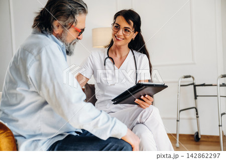 Female healthcare professional in white scrubs discusses treatment options with elderly male patient seated in a well-lit room with a walker nearby Female healthcare professional in white scrubs discusses treatment options with elderly male patient seated in a well-lit room with a walker nearby 142862297
