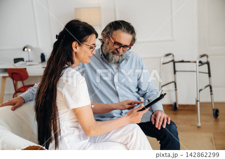 Female healthcare professional in white uniform discusses treatment options with elderly male patient in a well-lit room with modern decor and mobility aid nearby 142862299