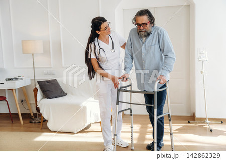 Female healthcare professional assists elderly man using walker in bright, modern living room with white walls and wooden floor, showcasing supportive care environment Female healthcare professional assists elderly man using walker in bright, modern living room with white walls and wooden floor, showcasing supportive care environment 142862329