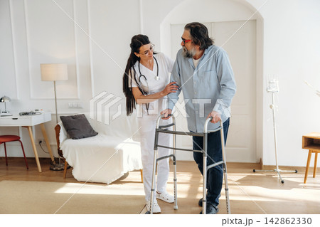 Female healthcare worker assists elderly man using walker in bright, modern living room with medical equipment and furniture in the background Female healthcare worker assists elderly man using walker in bright, modern living room with medical equipment and furniture in the background 142862330
