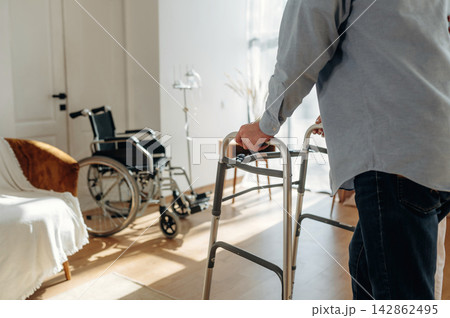 Elderly man using walker in a bright living room with a wheelchair and comfortable seating area visible in the background Elderly man using walker in a bright living room with a wheelchair and comfortable seating area visible in the background 142862495