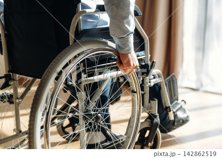 Individual in a wheelchair grips the wheel with one hand while seated indoors, sunlight streaming through a window illuminating the scene 142862519