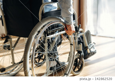 Individual in gray long-sleeve shirt grips the wheel of a manual wheelchair on wooden floor near large window with natural light streaming in Individual in gray long-sleeve shirt grips the wheel of a manual wheelchair on wooden floor near large window with natural light streaming in 142862520