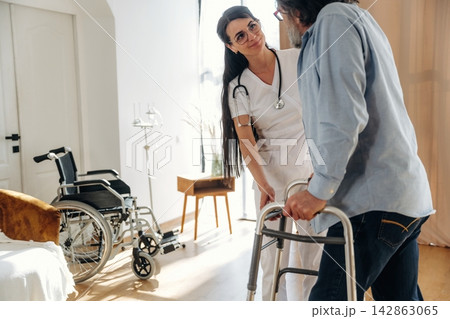 Female healthcare worker assists elderly man with walker in a bright room featuring a wheelchair and medical equipment in the background Female healthcare worker assists elderly man with walker in a bright room featuring a wheelchair and medical equipment in the background 142863065