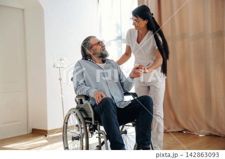 Female caregiver assists elderly man in wheelchair during a health check in a bright room with large windows and soft curtains, promoting comfort and care Female caregiver assists elderly man in wheelchair during a health check in a bright room with large windows and soft curtains, promoting comfort and care 142863093