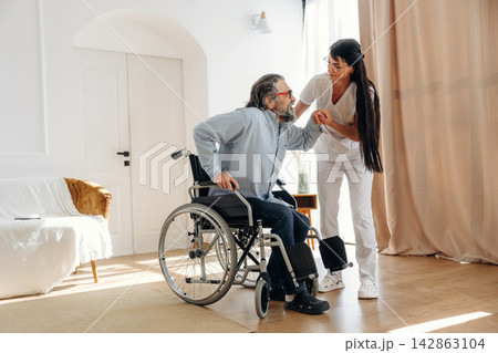 Elderly man in wheelchair assisted by caregiver in white uniform inside a bright, modern living room with wooden flooring and large windows Elderly man in wheelchair assisted by caregiver in white uniform inside a bright, modern living room with wooden flooring and large windows 142863104
