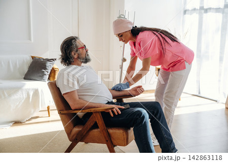 Female healthcare worker in pink scrubs measures blood pressure of elderly man in casual attire seated in a bright, modern living room with natural light Female healthcare worker in pink scrubs measures blood pressure of elderly man in casual attire seated in a bright, modern living room with natural light 142863118