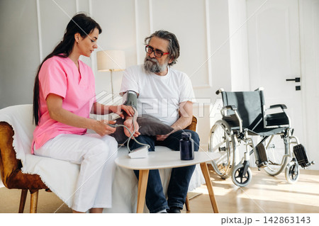 Female nurse in pink scrubs measures blood pressure of elderly man in white t-shirt seated on chair with wheelchair visible in bright, modern healthcare setting 142863143