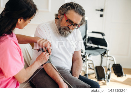Healthcare professional in pink scrubs measures blood pressure of elderly man with gray beard sitting on couch in home setting with wheelchair visible in the background Healthcare professional in pink scrubs measures blood pressure of elderly man with gray beard sitting on couch in home setting with wheelchair visible in the background 142863264