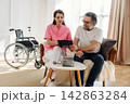 Female caregiver in pink scrubs assists elderly man with tablet in a cozy living room, wheelchair positioned nearby, sunlight streaming through large windows 142863284