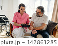 Female healthcare professional in pink scrubs discusses treatment plan with elderly male patient in living room with wheelchair and natural light 142863285