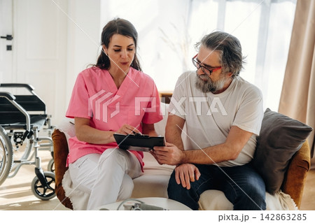 Female healthcare professional in pink scrubs discusses treatment plan with elderly male patient in living room with wheelchair and natural light 142863285
