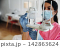 Female healthcare professional in pink scrubs adjusts IV drip with clear fluid in a medical facility, bright interior with furniture visible in the background 142863624