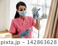 Female healthcare worker in pink scrubs adjusts IV drip stand while wearing gloves and a mask in a well-lit medical facility with large windows 142863628