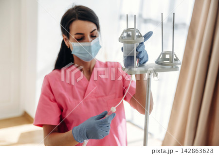 Female healthcare worker in pink scrubs adjusts IV drip stand while wearing gloves and a mask in a well-lit medical facility with large windows 142863628