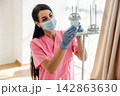 Female healthcare worker in pink scrubs adjusts IV drip stand while wearing a mask and gloves, bright hospital room with natural light and curtains in the background 142863630