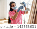 Female healthcare worker in pink scrubs adjusts IV drip in a bright hospital room with large windows and soft natural light illuminating the space 142863631