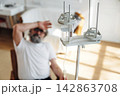 Elderly man with gray beard sitting in a chair, holding his forehead in discomfort, with an IV drip stand and medical equipment in a bright hospital room 142863708