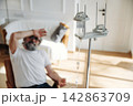 Elderly man with gray beard sitting in chair, resting hand on forehead, IV drip stand beside him in bright room with soft furnishings and natural light 142863709