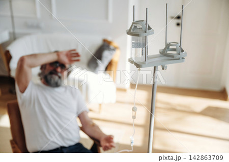 Elderly man with gray beard sitting in chair, resting hand on forehead, IV drip stand beside him in bright room with soft furnishings and natural light Elderly man with gray beard sitting in chair, resting hand on forehead, IV drip stand beside him in bright room with soft furnishings and natural light 142863709
