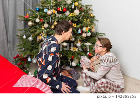 A couple sits near a Christmas tree on Christmas Day. They are exchanging gifts and enjoying the holiday spirit. The room has decorations and a festive atmosphere 142865258