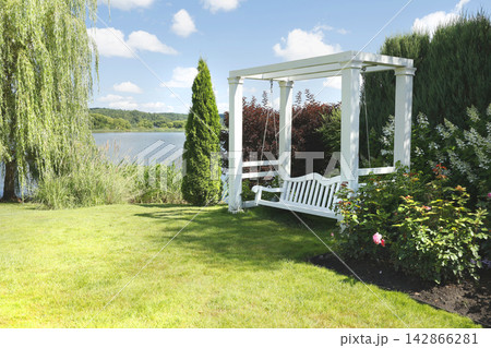 Elegant white wooden swing bench under pergola on green lakeside lawn. Luxury backyard relaxation zone featuring scenic waterfront view and beautiful landscape design. Contemporary garden decoration. Elegant white wooden swing bench under pergola on green lakeside lawn. Luxury backyard relaxation zone featuring scenic waterfront view and beautiful landscape design. Contemporary garden decoration. 142866281