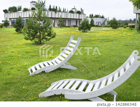 White wooden sun loungers on lush eco hotel lawn. Creative sustainable resort architecture landscaping design. Peaceful summer park garden hospitality relaxation nature background. White wooden sun loungers on lush eco hotel lawn. Creative sustainable resort architecture landscaping design. Peaceful summer park garden hospitality relaxation nature background. 142866348