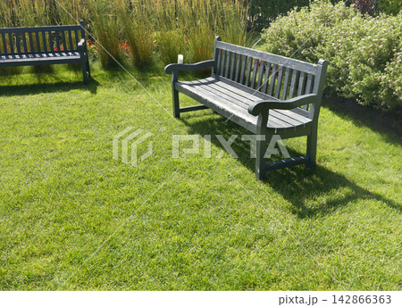Wooden bench on sunny green lawn. Elegant sustainable backyard patio seating area design. Peaceful summer park garden nature background. Wooden bench on sunny green lawn. Elegant sustainable backyard patio seating area design. Peaceful summer park garden nature background. 142866363