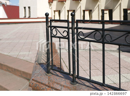 Forged iron railing granite stairs terrace. Vintage metal decorative fence on polished red granite step with blurred pavement and historic building facade background. Forged iron railing granite stairs terrace. Vintage metal decorative fence on polished red granite step with blurred pavement and historic building facade background. 142866697