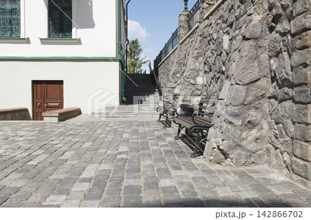 Restored European old town street cobbled pavement background. Traditional vintage wooden park benches stand near ancient stone masonry wall with stairs leading up to historic castle terrace facade. Restored European old town street cobbled pavement background. Traditional vintage wooden park benches stand near ancient stone masonry wall with stairs leading up to historic castle terrace facade. 142866702