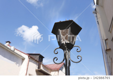 Vintage forged iron street lantern photo. Retro outdoor lamp post with decorated glass panes under blue sky. Antique classical city architecture element low angle view daylight. Vintage forged iron street lantern photo. Retro outdoor lamp post with decorated glass panes under blue sky. Antique classical city architecture element low angle view daylight. 142866765