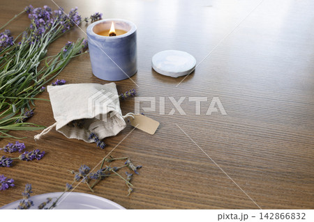 Open cotton sachet filled with dried lavender flowers on table. Scented candle burning near fabric pouch with blank craft tag. Cosy aromatherapy workshop background with large copy space. Open cotton sachet filled with dried lavender flowers on table. Scented candle burning near fabric pouch with blank craft tag. Cosy aromatherapy workshop background with large copy space. 142866832